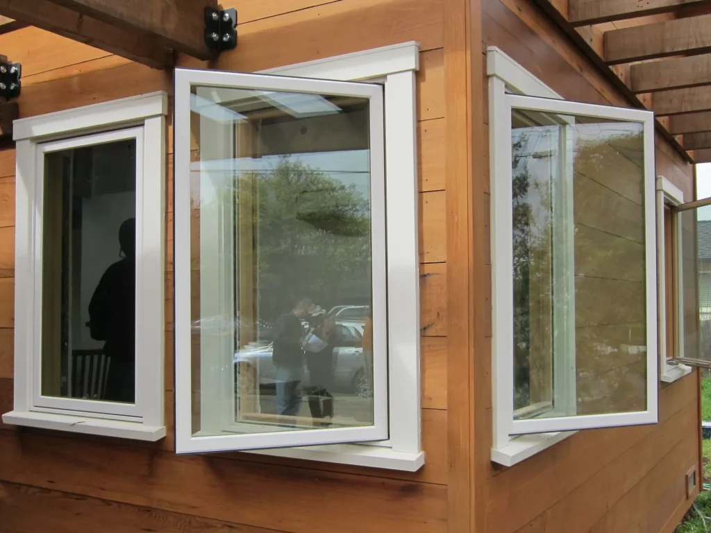 White framed casement windows open outward on a wooden exterior wall under a pergola